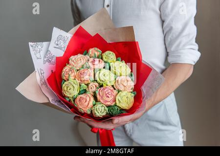 Ein gesichtsloser Mann hält einen Strauß süßer Marshmallow-Blumen in den Händen. Ein Geschenk in rotem Umschlag mit zarten Zuckerrosen auf hellem Hintergrund. Klein Stockfoto