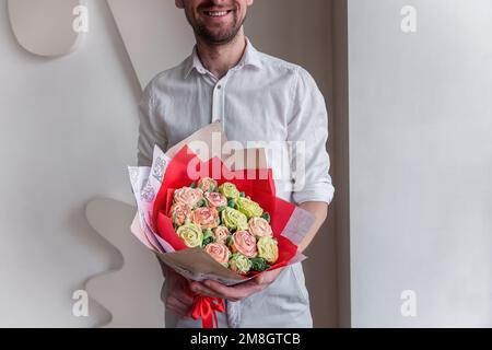 Ein gesichtsloser Mann hält einen Strauß süßer Marshmallow-Blumen in den Händen. Ein Geschenk in rotem Umschlag mit zarten Zuckerrosen auf hellem Hintergrund. Klein Stockfoto