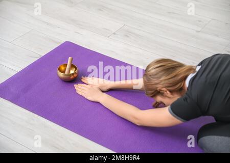 Junge Frau mit tibetischer Gesangsschale, die zu Hause Yoga praktiziert Stockfoto