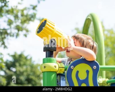 Das kleine Kind schaut in ein Spielzeugteleskop auf dem Spielplatz im Freien. Freizeitangebote für neugierige Kinder. Sommerspaß. Stockfoto