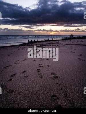 Fußabdrücke auf dem Sand führen zur Silhouette von zwei Personen bei Sonnenuntergang am Worthing Beach. Stockfoto