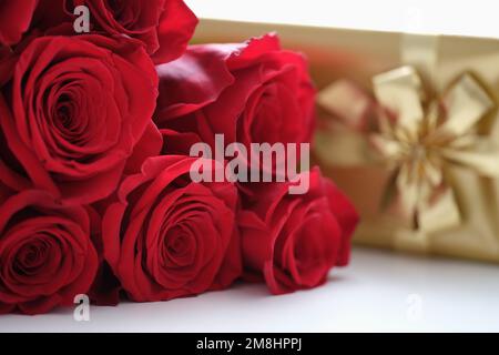 Frische rote Rosen und goldene Geschenkbox auf weißem Tisch Stockfoto