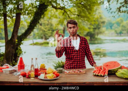 Ein Mann bereitet ein köstliches Abendessen für seine Freunde zu, die Spaß am Fluss in der Natur haben Stockfoto