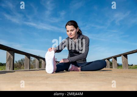 Sportliche Frau, die im Sitzen sitzt und sich vor dem Laufen oder Joggen ein Bein dehnt. Stockfoto