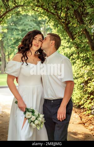 Wunderschöne, weiße, junge Paare spazieren im Garten. Schwarzhaariges Mädchen in festlichem Kleid und mit einem Strauß weißer Blumen Stockfoto