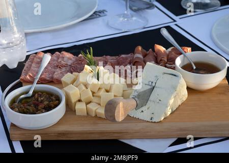 Auswahl an Käse und verarbeitetem Fleisch auf einem Holzteller in einem Restaurant im Freien mit Tageslicht und realistischer Komposition in einem Stockfoto