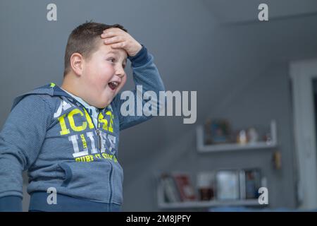 Der Junge steht, hält seinen Kopf mit einer Hand und sieht überraschend auf den Fernsehbildschirm, weil er endlich eine Mission erfolgreich abgeschlossen hat Stockfoto