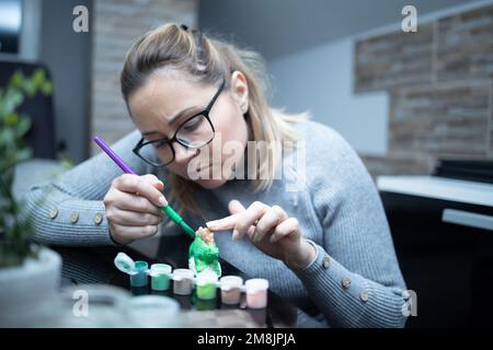 Eine Frau malt eine Keramikfigur eines Dinosauriers. Sie hält die Figur mit ihrem Finger. Der Pinsel hält sie in der anderen Hand. Er sieht konzentriert aus Stockfoto