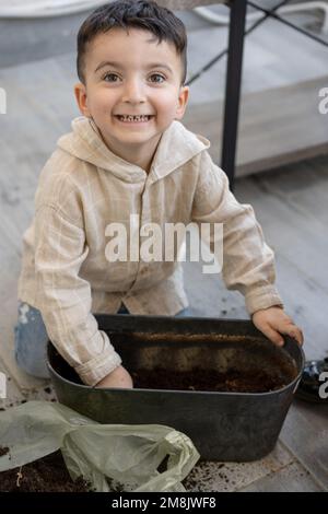 Komischer Junge, der zu Hause sitzt. Kleine Kinderschaufeln, Erde in Blumentopf geben. Stockfoto