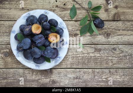 Frische blaue Pflaumen in Schüssel auf Holztisch mit Blättern und Zweigen, halbierte Pflaumenfrucht mit Samen als Dekoration, Draufsicht. Stockfoto