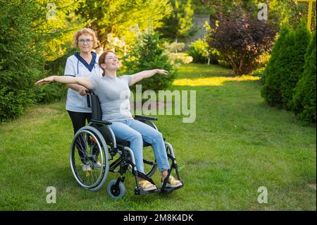 Eine ältere Krankenschwester geht mit einer Frau mittleren Alters im Rollstuhl durch den Park. Das Mädchen spreizte ihre Arme wie Flügel. Stockfoto