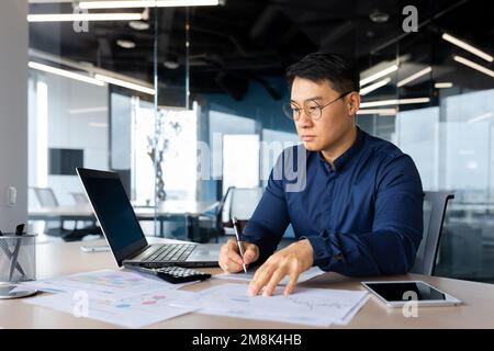 Konzentrierter junger Geschäftsmann Asiate arbeitet mit einem Laptop im Büro, schreibt Dokumente, Diagramme, Vereinbarungen, Rechnungen. Stockfoto