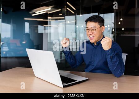 Ein glücklicher junger asiatischer Student sitzt im Büro am Tisch, sieht sich den Laptop an und freut sich. Die Prüfung bestanden, einen Brief erhalten, einen Job angenommen, die Universität betreten. Schreit vor Freude. Stockfoto