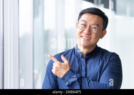 Nahaufnahme. Porträt eines jungen asiatischen Lehrers. Es steht im Büro, auf dem Campus in der Nähe des Fensters. Er sieht in die Kamera, lächelt, macht eine Erfolgsgeste mit dem Finger. Stockfoto