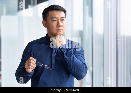 Porträt eines jungen asiatischen Lehrers. Er steht im Büro, auf dem Campus, hält eine Brille, stützt seinen Kopf mit der Hand, schaut durch das Fenster Stockfoto