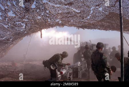Rückansicht links, Kamera unter Tarnnetz. Ein M198 155 mm Howitzer, Mündungsfeuer und Rauch. Bewaffnete Crewmitglieder im Vordergrund. Land: Unbekannt Stockfoto