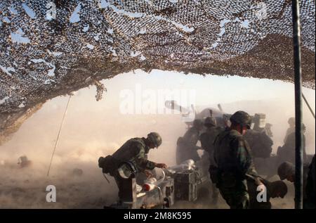 Rückansicht links, Kamera unter Tarnnetz. Ein M198 155 mm Howitzer, Mündungsfeuer und Rauch. Bewaffnete Crewmitglieder im Vordergrund. Land: Unbekannt Stockfoto