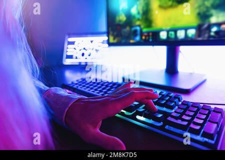 Die Hände des Spielers auf der Tastatur, aktives Drücken von Tasten, Spielen von MMO Games Online. Der Hintergrund wird mit Neonlichtern beleuchtet. Stockfoto