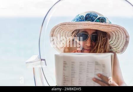 Porträt einer Frau mit Hut, die sich auf einen durchsichtigen Stuhl lehnt und eine Zeitschrift liest. Das blaue Meer im Hintergrund. Urlaubskonzept Stockfoto