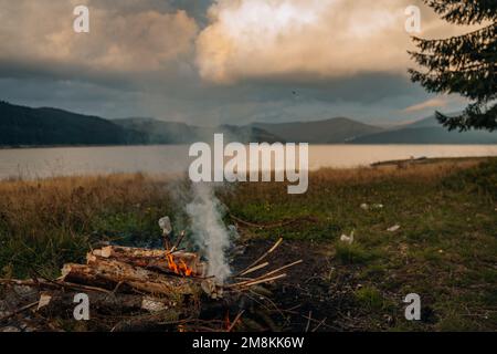 Ein kleines Lagerfeuer mit sanften Flammen neben einem See während eines glühenden Sonnenuntergangs Stockfoto