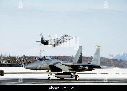 Eine F-15 startet als eine andere, die zur Vorbereitung auf den Start auf der schneefreien Piste 05 fährt. Basis: Luftwaffenstützpunkt Elmendorf Bundesstaat: Alaska (AK) Land: Vereinigte Staaten von Amerika (USA) Stockfoto
