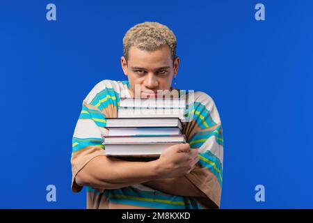 Fauler Schüler ist unzufrieden mit der Menge an Büchern, Hausaufgaben in der Highschool, blauer Hintergrund. Ein verärgerter Kerl ist verärgert, entmutigt Stockfoto