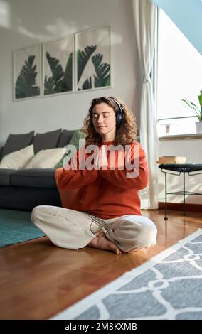 Eine junge, aufmerksame Frau mit Kopfhörern, die zu Hause meditiert. Vertikal Stockfoto