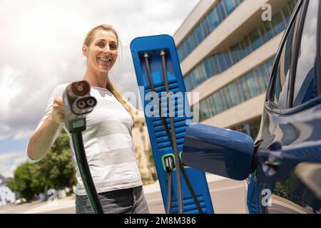 Eine Frau hält einen DC CCS2 EV-Ladeanschluss am Hypercharger oder an der Supercharger-Ladestation, um sie an die Steckdose ihres Autos anzuschließen. Stockfoto