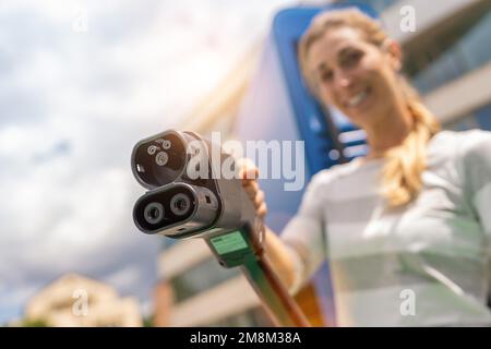 Glückliche Frau, die einen DC CCS2 EV-Ladeanschluss am Hypercharger oder Supercharger hält, um ihr Auto aufzuladen. Stockfoto