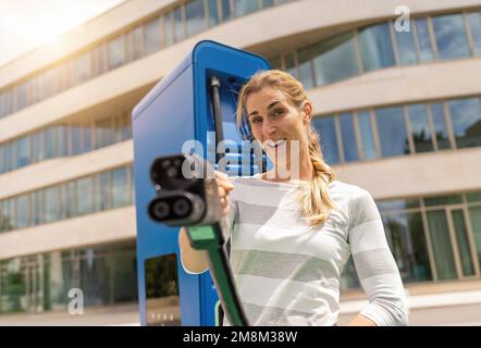 Frau hält einen DC CCS2 EV-Ladeanschluss an einem Hypercharger oder Supercharger, um ihr Auto aufzuladen. Stockfoto