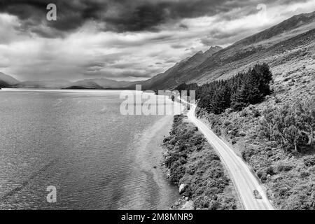 Dramatischer, kontrastreicher schwarzweißer Blick auf das Auto auf dem Highway 6 entlang des Lake Wkatipu in Otago von Neuseeland - malerische Luftlandschaft. Stockfoto