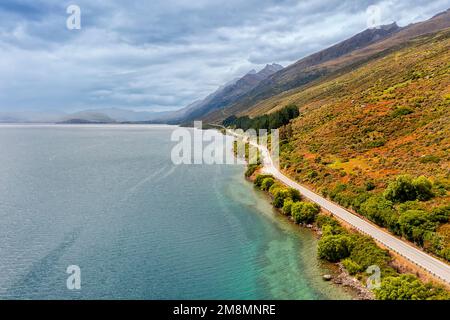 Abgelegene Autobahn entlang des Wakatipu Sees auf der Südinsel Neuseelands unter malerischen Bergketten an einem bewölkten sonnigen Tag. Stockfoto