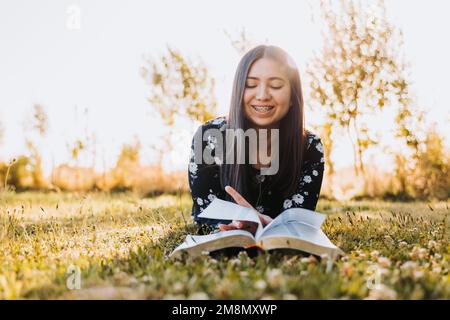 Lächelndes junges, glaubhaftes Mädchen, das auf dem Gras lag und ihre bibel studierte, auf dem Feld bei Sonnenuntergang. Speicherplatz kopieren Stockfoto