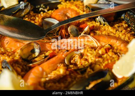 Traditionelle Paella im Restaurant, Meeresfrüchte-Paella in der Pfanne mit einem riesigen Eisenlöffel, Garnelen und Muscheln, Zitrone Stockfoto