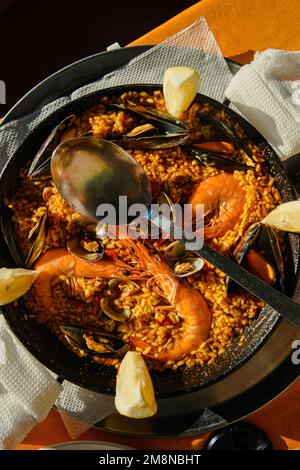 Traditionelle Paella im Restaurant, Meeresfrüchte-Paella in der Pfanne mit einem riesigen Eisenlöffel, Garnelen und Muscheln, Zitrone Stockfoto