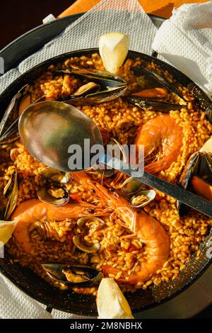 Traditionelle Paella im Restaurant, Meeresfrüchte-Paella in der Pfanne mit einem riesigen Eisenlöffel, Garnelen und Muscheln, Zitrone Stockfoto