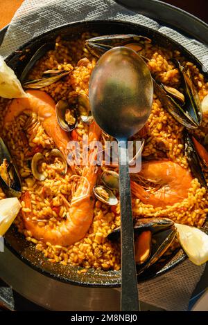 Traditionelle Paella im Restaurant, Meeresfrüchte-Paella in der Pfanne mit einem riesigen Eisenlöffel, Garnelen und Muscheln, Zitrone Stockfoto