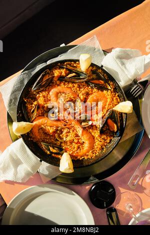 Traditionelle Paella im Restaurant, Meeresfrüchte-Paella in der Pfanne mit einem riesigen Eisenlöffel, Garnelen und Muscheln, Zitrone Stockfoto