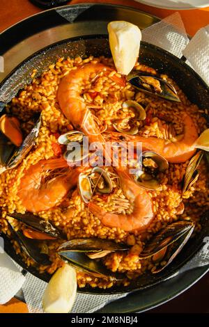 Traditionelle Paella im Restaurant, Meeresfrüchte-Paella in der Pfanne mit einem riesigen Eisenlöffel, Garnelen und Muscheln, Zitrone Stockfoto