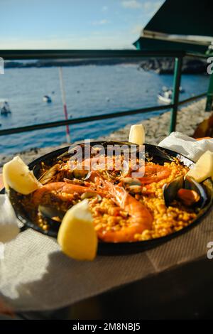 Paella mit einem riesigen Eisenteller, Garnelen und Muscheln, Zitrone. Zum Hintergrund des Meeres in Spanien im Sommer. Stockfoto