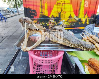 Krokodilfleisch zum Verkauf, aufgespießt auf Bambusstäbchen, die getrockneten Köpfe auf einem Silbertablett, benutzt, um für das beliebte thailändische Fleisch zu werben, zum Verkauf in der Nähe eines Tempels Stockfoto