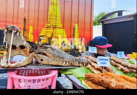 Krokodilfleisch zum Verkauf, aufgespießt auf Bambusstäbchen, die getrockneten Köpfe auf einem Silbertablett, benutzt, um für das beliebte thailändische Fleisch zu werben, zum Verkauf in der Nähe eines Tempels Stockfoto