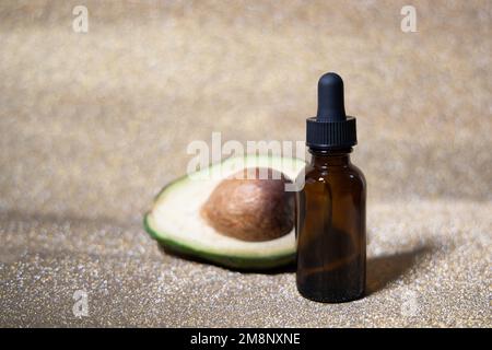 Avocadoöl. Ätherisches Öl in Glasflasche auf glänzendem goldenen Hintergrund. Avocado für gesunde Haut und Haare. Kosmetikinridient. Platz für Text. Stockfoto