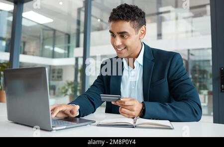 Junger, glücklicher Geschäftsmann, der eine Kreditkarte in der Hand hat und benutzt und auf einem Laptop tippt, allein auf der Arbeit. Ein hispanischer Geschäftsmann, der lächelt, während er einen macht Stockfoto