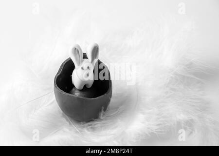 Weißer Osterhasen in einem zerbrochenen Schokoladenei in einem weißen Federnest, schwarz-weiß. Süße Ostertradition. Frühlingsferien. Ostergeschenk für Stockfoto