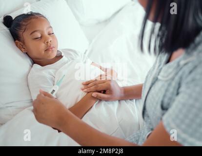 Krankes kleines Mädchen im Bett, während ihre Mutter ein Thermometer benutzt, um ihre Temperatur zu messen. Junger besorgter alleinerziehender Elternteil, der mit einem kranken Kind sitzt, während sie sich Stockfoto