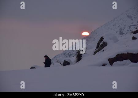 Srinagar, Indien. 14. Januar 2023. Ein Bewohner benutzt sein Handy auf dem schneebedeckten Hügel während des Sonnenuntergangs am Stadtrand von Srinagar. Das Wetter verbesserte sich im Kaschmir-Tal nach Tagen mit moderatem bis starkem Schneefall und Regen. In der Zwischenzeit wurden am Samstag Lawinen im Bezirk Bandipora und im Bahnhof Sonamarg gemeldet, bei denen es in den letzten Tagen zu moderaten bis schweren Schneefällen kam. Dies ist der zweite Vorfall, der in den letzten zwei Tagen in der beliebten Hill Station gemeldet wurde. Zwei Arbeiter starben am Donnerstag, als eine Lawine die Baustelle einer Baufirma in Sonamarg traf. Kredit: Stockfoto