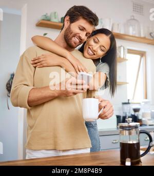 Glückliches, junges gemischtrassiges Paar, das zu Hause liebevoll und liebevoll ist. Junger Kaukasier, der sein Smartphone benutzt und Kaffee trinkt, während seine Freundin Stockfoto