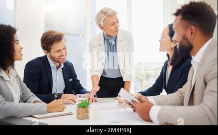 Eine reife kaukasische Geschäftsfrau, die während eines Meetings in einem Sitzungssaal eine Präsentation mit ihren verschiedenen Kollegen führt. Geschäftsleute gehen Stockfoto