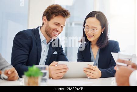 Zwei glückliche junge, vielfältige Kollegen diskutieren Pläne und Ideen auf einem digitalen Tablet-Gerät während einer Besprechung in einer Vorstandssitzung. Asiatisch Stockfoto
